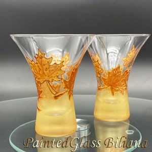 May include: Two clear glass vases with a flared top and a golden-yellow base. The vases are decorated with hand-painted orange and gold autumn leaves. The text "Painted Glass Biliana" is at the bottom of the image.