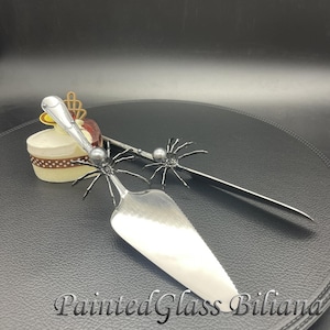 May include: A silver cake knife and server set with black spider accents and pearl-like beads. The set is on a black surface.