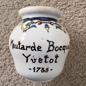May include: White ceramic jar with a rounded body and a narrow neck. The jar features the text "Moutarde Bocquel Yvetot -1735" in dark brown script. The rim is white, and the jar is decorated with blue, yellow, and green floral patterns.