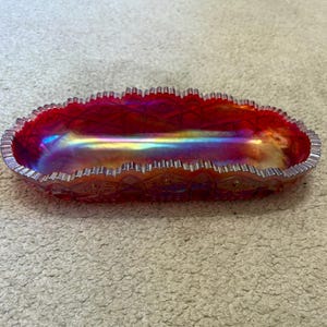 May include: An oval-shaped carnival glass dish featuring a vibrant red hue with an iridescent, multi-colored sheen. The dish has a scalloped, serrated edge and a textured pattern along the exterior sides, creating a reflective surface.