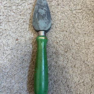 May include: A vintage cheese knife with a green wooden handle and a dull, gray blade. The blade has a teardrop shape. The handle is painted green and shows signs of wear. The knife is designed for cutting cheese.