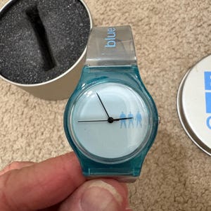 Puede incluir: Un reloj de pulsera azul turquesa con esfera azul claro y manecillas negras. La esfera del reloj presenta una silueta de tres figuras. La correa del reloj es transparente con la palabra "blue" impresa. El reloj est&aacute; en su embalaje original.