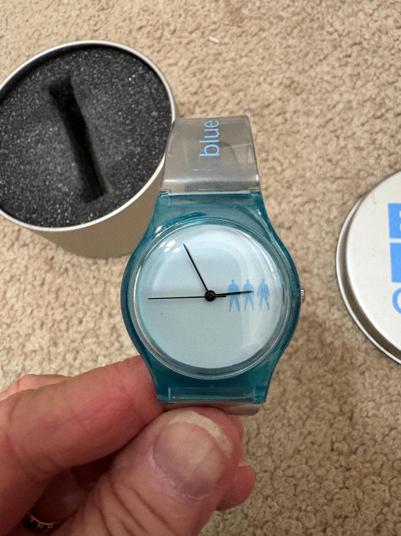 Puede incluir: Un reloj de pulsera azul turquesa con esfera azul claro y manecillas negras. La esfera del reloj presenta una silueta de tres figuras. La correa del reloj es transparente con la palabra "blue" impresa. El reloj est&aacute; en su embalaje original.