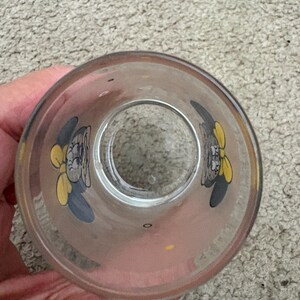 May include: A clear glass tumbler featuring a repeating graphic print of a cartoon character with grey ears and a yellow bow. The design is positioned around the interior wall of the glass, which has a circular opening at the top and base.