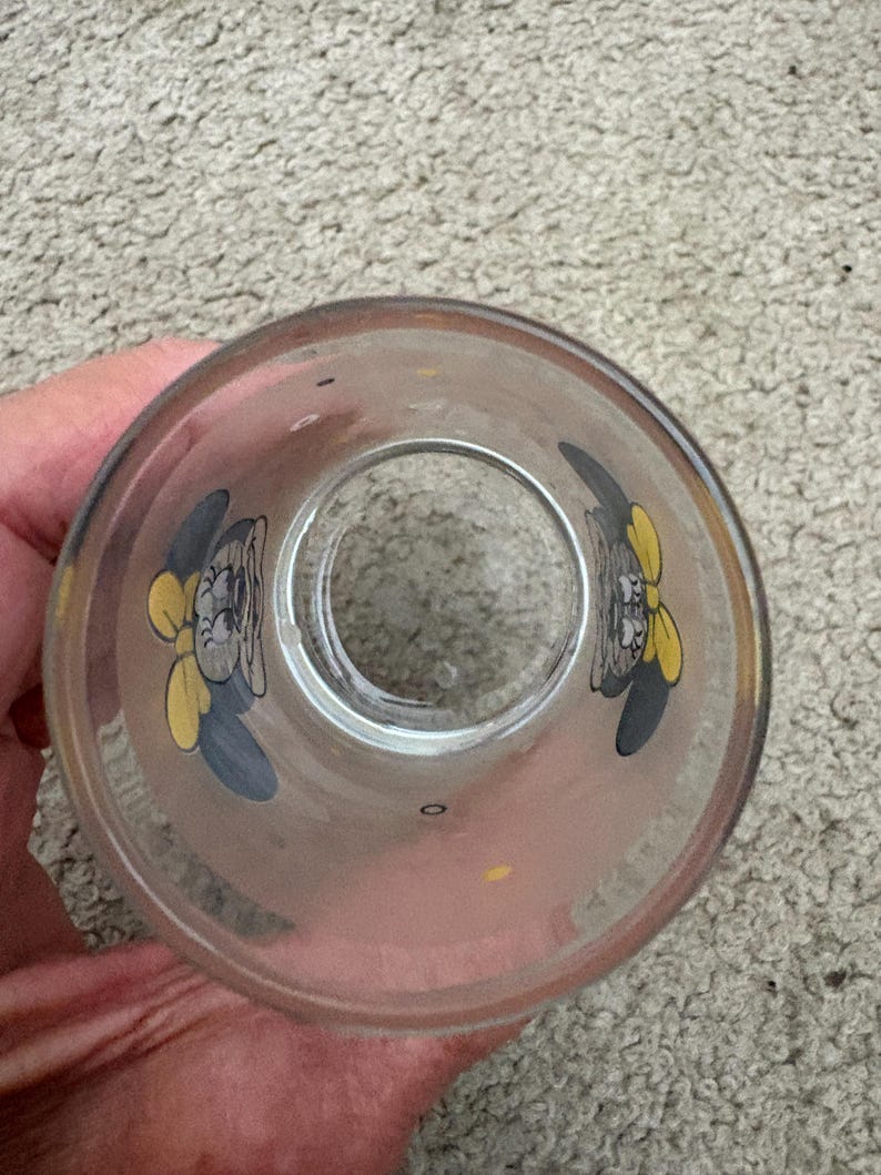 May include: A clear glass tumbler featuring a repeating graphic print of a cartoon character with grey ears and a yellow bow. The design is positioned around the interior wall of the glass, which has a circular opening at the top and base.