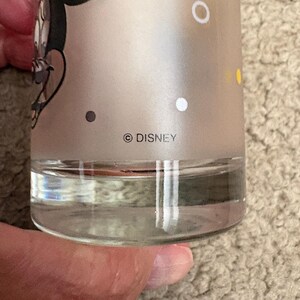 May include: A frosted glass tumbler featuring a graphic of Minnie Mouse with a yellow bow. The glass has a clear base and includes white and yellow polka dots printed on the surface. The text DISNEY is printed in black near the bottom edge.