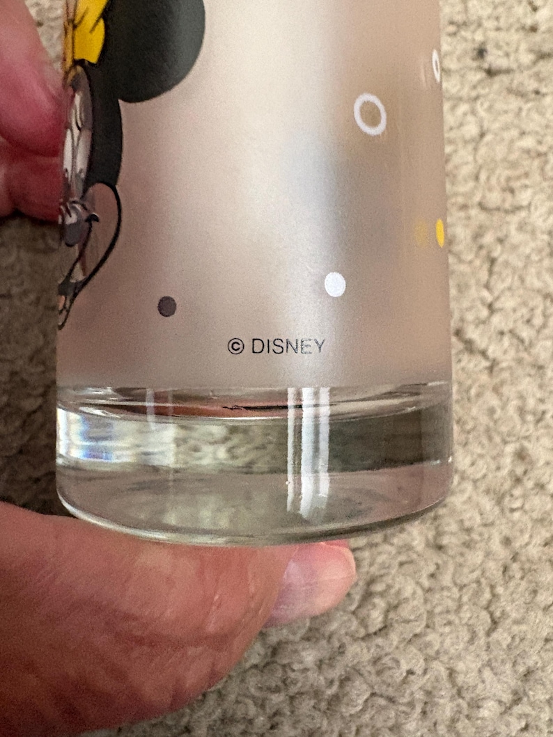 May include: A frosted glass tumbler featuring a graphic of Minnie Mouse with a yellow bow. The glass has a clear base and includes white and yellow polka dots printed on the surface. The text DISNEY is printed in black near the bottom edge.