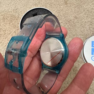 Puede incluir: Un reloj de pulsera transparente con detalles en azul. La esfera del reloj es plateada con una l&iacute;nea roja. El reloj se sostiene en una mano, con un anillo negro visible. El reloj est&aacute; en su embalaje original.