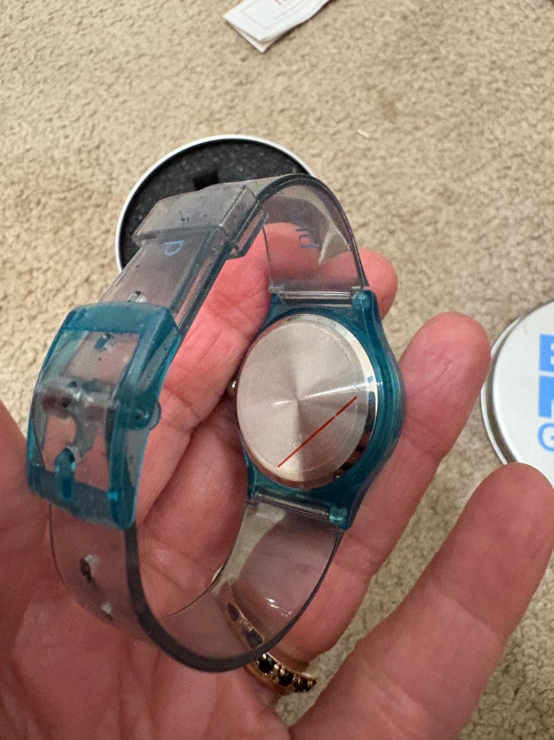 Puede incluir: Un reloj de pulsera transparente con detalles en azul. La esfera del reloj es plateada con una l&iacute;nea roja. El reloj se sostiene en una mano, con un anillo negro visible. El reloj est&aacute; en su embalaje original.
