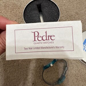 Puede incluir: Un reloj de cuarzo turquesa y plateado con una correa a juego se muestra junto a una tarjeta de garant&iacute;a y una caja de reloj. La tarjeta dice "Pedre Quartz Watches" y "Two Year Limited Manufacturer's Warranty".