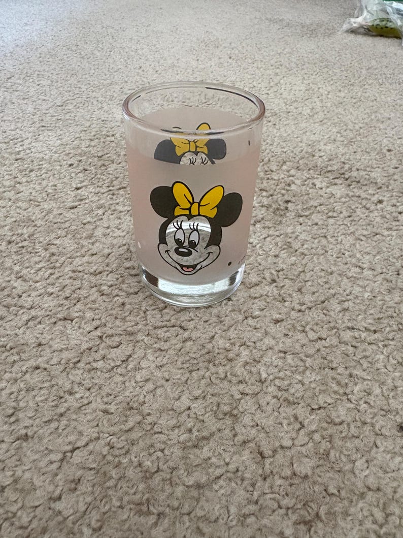 May include: A clear glass tumbler featuring a printed graphic of Minnie Mouse with a yellow bow. The glass has a slightly frosted appearance and a cylindrical shape with a thick, transparent base, showcasing the character design on the front.