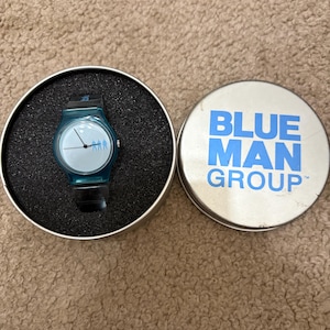 Puede incluir: Un reloj de pulsera turquesa con una correa negra se encuentra en un estuche redondo negro. La esfera del reloj tiene un fondo azul claro con tres peque&ntilde;as figuras. Junto a &eacute;l, una lata plateada con las palabras "BLUE MAN GROUP" en azul.