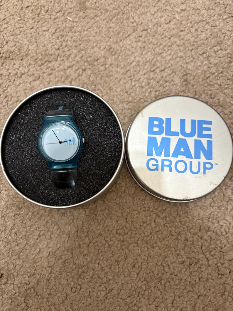 Puede incluir: Un reloj de pulsera turquesa con una correa negra se encuentra en un estuche redondo negro. La esfera del reloj tiene un fondo azul claro con tres peque&ntilde;as figuras. Junto a &eacute;l, una lata plateada con las palabras "BLUE MAN GROUP" en azul.