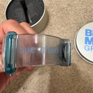 Puede incluir: Un recipiente transparente te&ntilde;ido de azul para productos de aseo con las palabras "blue man" impresas en el lateral. Junto a &eacute;l, un recipiente plateado con una esponja negra y una tapa azul y blanca con las palabras "BLUE MAN GROOMING".