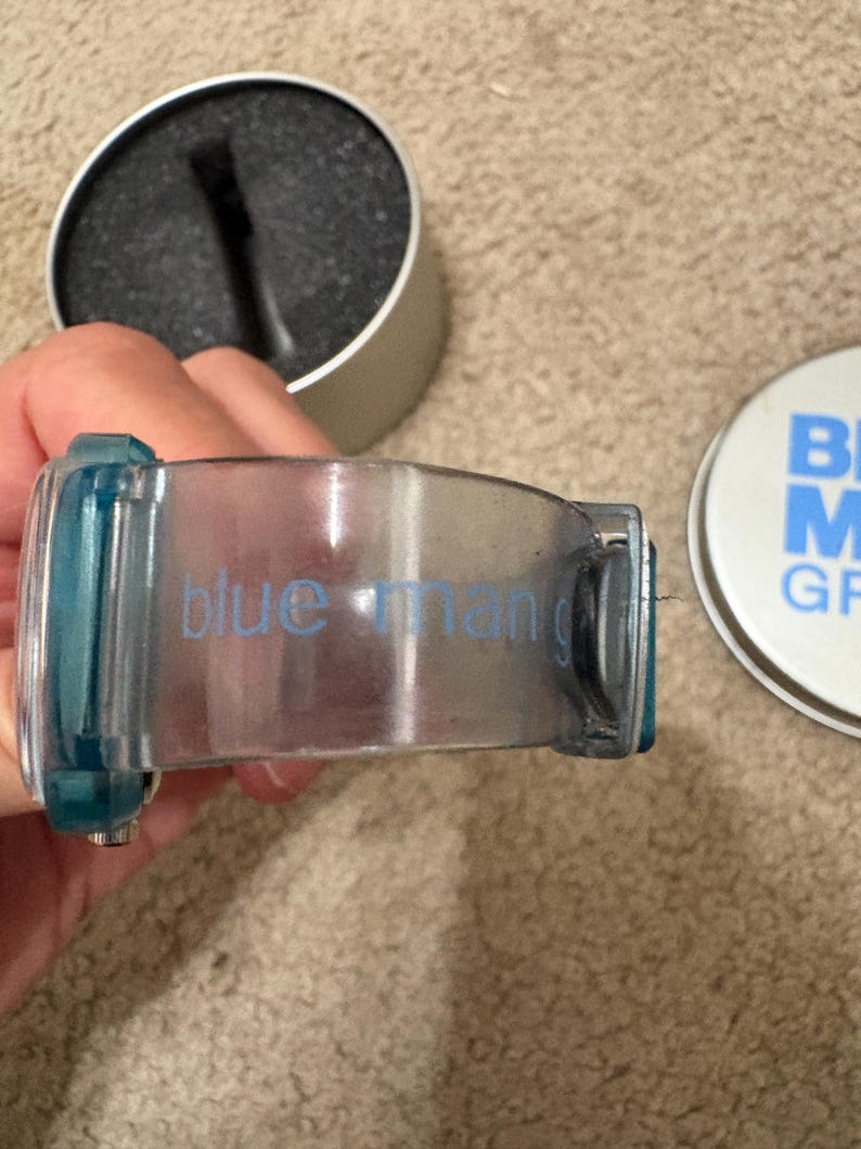 Puede incluir: Un recipiente transparente te&ntilde;ido de azul para productos de aseo con las palabras "blue man" impresas en el lateral. Junto a &eacute;l, un recipiente plateado con una esponja negra y una tapa azul y blanca con las palabras "BLUE MAN GROOMING".