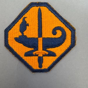 May include: Embroidered patch with a yellow and blue design. The patch features a stylized anvil, sword, and a small figure. The patch has a dark blue border and is in a unique octagonal shape.