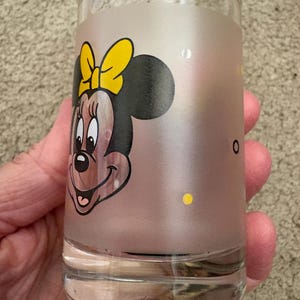 May include: A clear glass tumbler featuring a frosted middle section decorated with a graphic of Minnie Mouse wearing a yellow bow. The design includes small yellow polka dots scattered across the frosted surface, while the top and bottom rims remain clear.