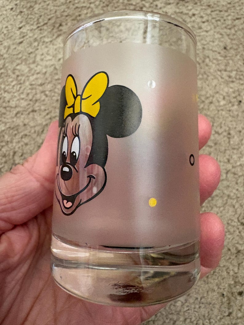 May include: A clear glass tumbler featuring a frosted middle section decorated with a graphic of Minnie Mouse wearing a yellow bow. The design includes small yellow polka dots scattered across the frosted surface, while the top and bottom rims remain clear.