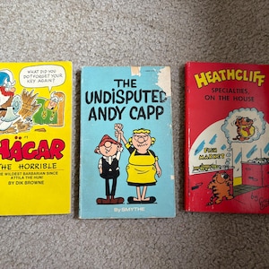 May include: Three vintage comic books. The first is yellow with a cartoon of a man with a beard and a helmet. The title is "Hagar the Horrible". The second is blue with the title "The Undisputed Andy Capp". The third is red with the title "Heathcliff Specialties, On the House".
