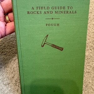 May include: A green book titled "A Field Guide to Rocks and Minerals" with the author's name, Pough, below the title. A brown illustration of a geological hammer is on the cover. The book is held by a hand.