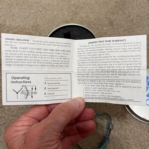 Puede incluir: Un reloj con una correa azul y una caja plateada se muestra con su garant&iacute;a e instrucciones de funcionamiento. El reloj es parcialmente visible, descansando sobre una superficie de color claro. La informaci&oacute;n de la garant&iacute;a est&aacute; impresa en texto negro.