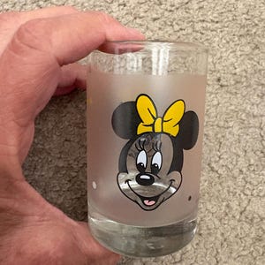 May include: A clear glass tumbler featuring a frosted finish and a graphic print of Minnie Mouse. The character is depicted with her signature black ears, a bright yellow bow, and a smiling expression. The glass has a smooth, cylindrical shape.