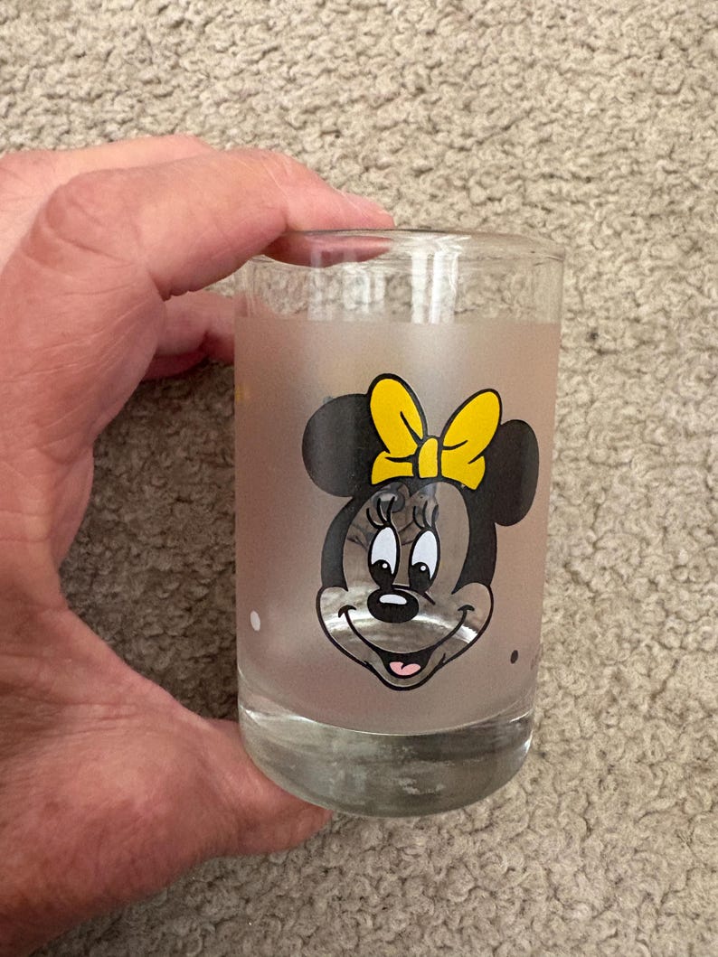 May include: A clear glass tumbler featuring a frosted finish and a graphic print of Minnie Mouse. The character is depicted with her signature black ears, a bright yellow bow, and a smiling expression. The glass has a smooth, cylindrical shape.