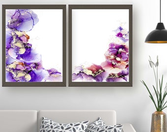 Puede incluir: Dos impresiones de arte abstracto enmarcadas con diseños de acuarela en tonos morados, rosas y dorados. Las impresiones están colgadas en una pared blanca sobre un sofá blanco con dos cojines de tiro de patrón gris.