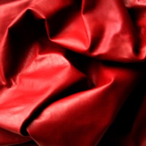 May include: Close-up of a vibrant red leather fabric with a glossy finish. The material is heavily draped, creating deep shadows and highlights that emphasize its texture and sheen. The folds and creases suggest a supple, pliable quality.