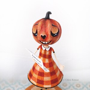 Peut inclure: Une figurine avec une tête de citrouille et un nez en forme de cœur, portant une robe à carreaux orange et jaune avec un col blanc. La figurine tient un papier blanc avec le texte CHANSON D'AUTOMNE et repose sur un socle rond orange étoilé.