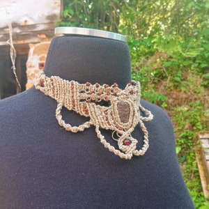 May include: A beige macrame choker necklace with brown and red accents. The necklace features a central rectangular stone and smaller red beads. The design includes intricate knotting and decorative loops, displayed on a black mannequin.