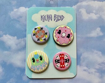Könnte beinhalten: Ein Set aus vier runden Emaille-Anstecknadeln auf einer hellblauen Karte mit dem Text "KIERRA BOYD". Die Anstecknadeln zeigen verschiedene Designs, darunter ein Pilzmuster, ein rosa Donut, ein Clown und eine rosa Anstecknadel mit Totenköpfen.