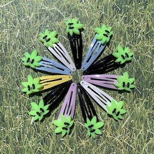 Puede incluir: Una colección de horquillas de colores dispuestas en círculo. Cada horquilla tiene un color diferente, incluyendo negro, blanco, amarillo y azul. Cada horquilla está adornada con un diseño de hoja verde de estilo caricatura con una cara.