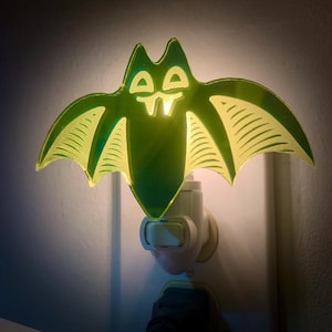 May include: A green bat-shaped night light plugged into a white wall outlet. The bat has a detailed wing design and a glowing center. The light is a decorative item, perfect for Halloween or themed decor.