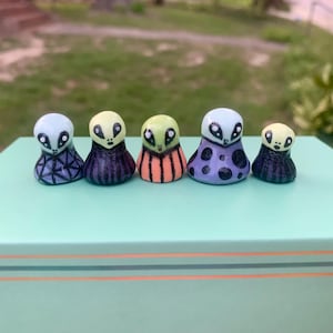 May include: Five small, rounded, hand-painted figurines shaped like alien heads with distinct patterns. The first has a blue geometric grid, the second has dark vertical stripes, the third has orange stripes, the fourth has purple polka dots, and the fifth has dark stripes.
