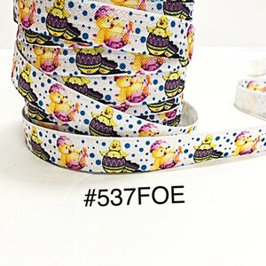 May include: White ribbon with a repeating pattern of yellow chicks in decorated Easter eggs, with blue polka dots. The ribbon is rolled up in a spool. The text "#537FOE" is printed on the ribbon.