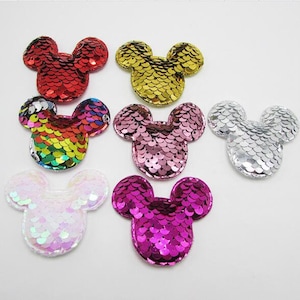 May include: A collection of sequined Mickey Mouse head patches in various colors, including red, gold, rainbow, pink, silver, white, and magenta. The patches are arranged on a white surface, showcasing their shiny, reflective surfaces.