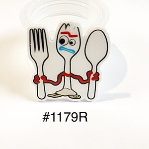 May include: A white plastic charm featuring a cartoon character resembling a spork with googly eyes, a red eyebrow, and a blue mouth. The character holds a white fork on the left and a white spoon on the right with red arms. The text #1179R is printed below.
