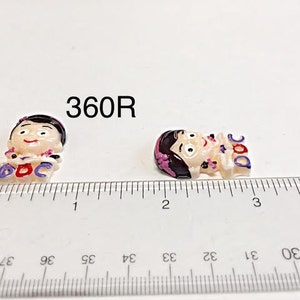 May include: Two small, flat, resin-like character charms featuring a cartoon figure with black hair and a pink headband. The figures have the word DOC printed in purple and blue letters. A clear plastic ruler shows the charms are about 1 inch wide.