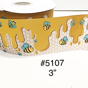 May include: A spool of yellow ribbon featuring a repeating pattern of cartoon bees and dripping honey over a white honeycomb background. The ribbon is labeled with the text #5107 3 inches, indicating the width of the material on the spool.