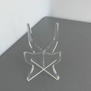 May include: Clear acrylic display stand with a curved top and an X-shaped base. The stand is designed to hold and showcase small items. The stand is made of clear acrylic and is suitable for displaying collectibles.