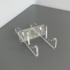 May include: A clear acrylic display stand featuring a rectangular back support and two forward-extending arms with notched ends. The stand is constructed from transparent material and is designed to hold items in an upright position for viewing.