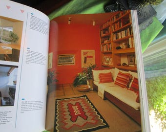 May include: An open book displaying interior design photography. The right page features a room with bright orange walls, a built-in wooden bookshelf filled with books, a long sofa with striped cushions, and a patterned rug on the floor.