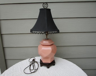 May include: A table lamp featuring a faceted, light pink ceramic base with a matching lid, set upon a dark brown wooden stand. It is topped with a black fabric lampshade adorned with hanging black beads and a decorative metal finial at the top.