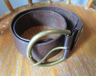 May include: A dark brown leather belt featuring a smooth, wide strap and a large, rounded brass buckle. The belt is coiled into a circular shape, showcasing the deep brown texture of the leather and the metallic finish of the brass hardware.