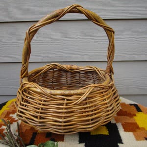 May include: A round, woven wicker basket with a handle. The basket is made of light brown wicker and has a sturdy handle that arches over the top. The basket is sitting on a colorful patterned surface.