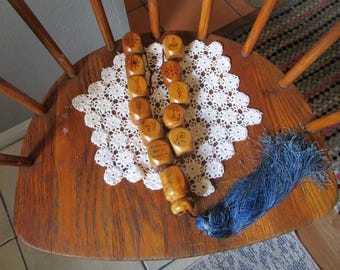 May include: A string of light brown wooden beads, each featuring a unique dark brown etched symbol, arranged in two parallel lines. The beads are connected by a dark cord and terminate in a long, textured blue tassel at the bottom end.