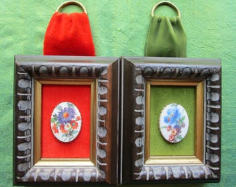 May include: Two small rectangular dark brown wooden frames with ornate edges. Each frame contains a velvet backing, one red and one green, with a gold-toned metal ring at the top. Centered in each is an oval porcelain plaque featuring floral art.