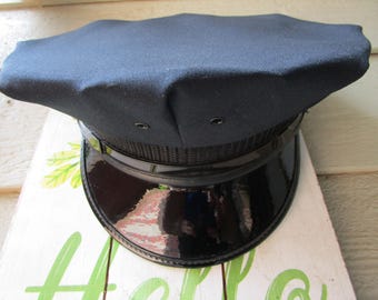 May include: A dark navy blue service cap featuring a structured flat top, a black textured band, and a glossy black patent leather visor. The cap includes two small metal ventilation eyelets on the front panel above the black band.