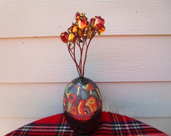 May include: A rounded ceramic vase with a dark base and a colorful upper section featuring abstract patterns in red, yellow, blue, and green. A bouquet of dried roses with yellow and red petals is arranged inside the narrow neck of the vase.